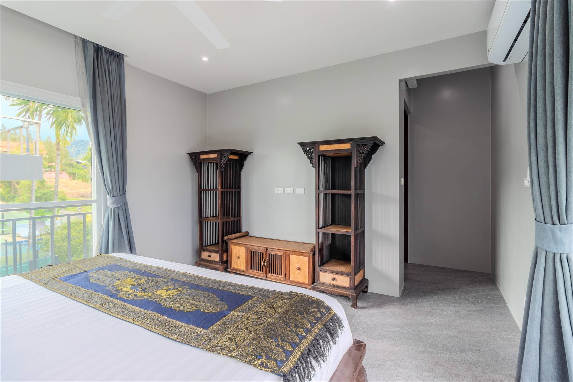 Spacious bedroom with Thai silk bed runner, carved wooden shelving, and pool-area views