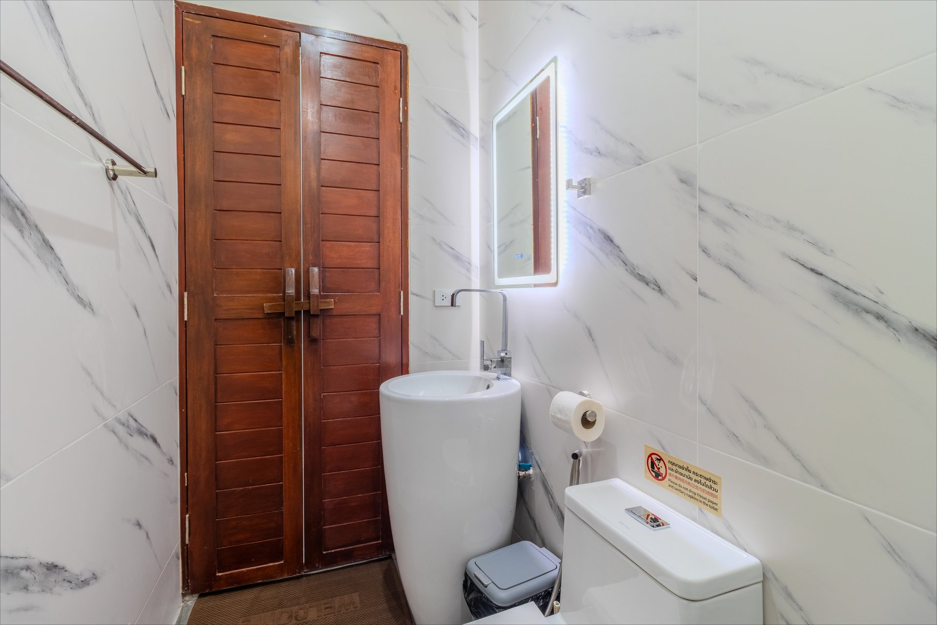Modern marble-walled bathroom with LED-lit mirror, pedestal basin, and teak wood doors